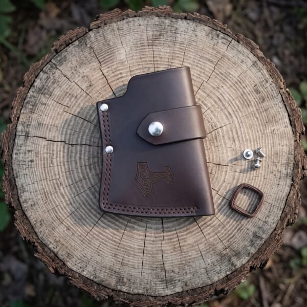 Leather axe sheath with a stamped Mjolnir and stitched welt seen from above on a cut log with spare snaps and a keeper