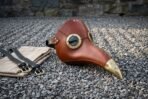 Leather plague doctor mask with brass rimmed goggles and brass beak cap lying on gravel next to its straps and bag
