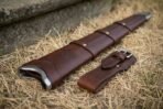 Leather sword scabbard with stamped Valknut at the steel throat lying on straw grass next to a separate belt frog