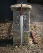 Long hand forged medieval arming sword with an engraved arched crossguard and tan leather grip leaning against a weathered stone gatepost beside a leather scabbard
