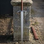 Long hand forged medieval arming sword with an engraved arched crossguard and tan leather grip leaning against a weathered stone gatepost beside a leather scabbard