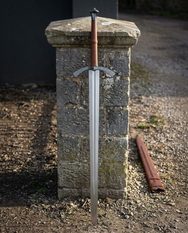 Long hand forged medieval arming sword with an engraved arched crossguard and tan leather grip leaning against a weathered stone gatepost beside a leather scabbard