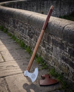 Long handled viking axe with a polished bearded head engraved with a Vegvisir, leather wrapped haft, leaning on a stone parapet beside a fitted leather blade cover