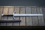 Long medieval arming sword in hand forged 1095 steel with a downswept crossguard and etched fuller panel laid across grey weathered decking boards