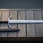 Long medieval arming sword in hand forged 1095 steel with a downswept crossguard and etched fuller panel laid across grey weathered decking boards