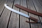 Long view down a hooked single edged blade with wooden grip and braided tan lanyard beside its scabbard on decking