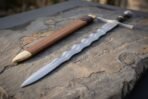 Long view down a wavy flamberge blade toward the disc pommel, wooden scabbard with brass chape alongside on slate