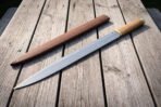 Long Viking seax blade with light wood grip shown full length on wooden planks, brown leather sheath laid parallel