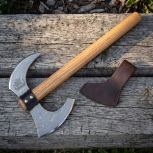 Longship damascus axe with a Norse ship under sail cut into the bearded head, hooked horn and riveted leather collar lying on grey weathered planks beside its blade mask