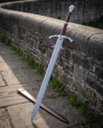 Longship etched damascus longsword with a barbed guard, twin parrying rings, burgundy leather grip, fluted crown pommel and leather scabbard against an old stone wall