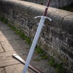 Longship etched damascus longsword with a barbed guard, twin parrying rings, burgundy leather grip, fluted crown pommel and leather scabbard against an old stone wall