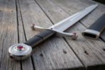 Low angle detail of a jewelled arming sword hilt on decking, black leather grip and disc pommel with a red glass cabochon set into the polished steel face