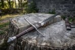Low angle of a flame bladed sword on a weathered stump with its scabbard and belt standing behind in dappled light