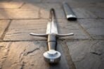 Low angle view down a steel arming sword from the wheel pommel and black grip, with the scabbard behind on paving