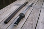 Low angle view of a ninjato sword on a weathered wooden table, four triple spirals engraved around the satin steel guard above a black cord wrapped grip