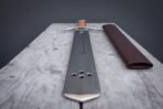 Low view along a steel falchion blade from the clipped tip with three holes toward the hilt, brown scabbard beside it