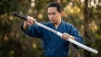 Man drawing a steel Japanese sword with black wrapped handle and round guard from a gloss white saya in a garden