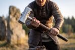 Man gripping a steel war hammer with runes on the head and a black leather grip beside standing stones