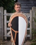 Man holding a black and white Norman kite shield with leather straps in front of a wooden farm gate