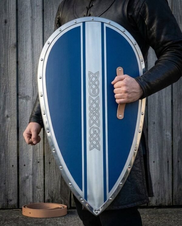 Man holding a blue Norman kite shield by its leather grip, riveted steel rim and knotwork band facing the camera