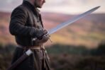 Man in a dark coat holding a steel basket hilt broadsword on a heather covered hillside with a scabbard at his hip