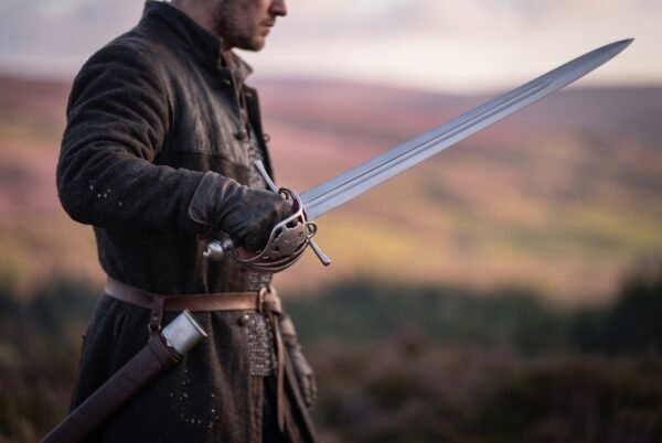 Man in a dark coat holding a steel basket hilt broadsword on a heather covered hillside with a scabbard at his hip