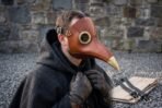 Man in a dark hooded cloak wearing a brown leather plague doctor mask with brass goggles beside a stone wall
