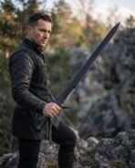 Man in a dark leather jacket holding a black broadsword with swept guard and ribbed grip among rocks and trees
