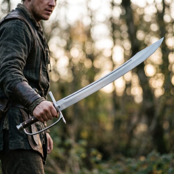 Man in a dark tunic and leather bracer holding a curved messer with knuckle bow and wood grip in a sunlit forest