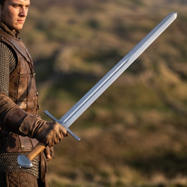 Man in mail and leather holding a blunt arming sword with straight cross guard and tan leather grip on a hillside