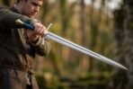 Man in a wool tunic holding a steel sword with blue leather grip and brass pommel out at arm's length in a forest