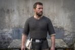 Man wearing a short sleeved blackened steel chainmail shirt with a leather belt and pouch against a weathered stone wall