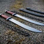 Matched chain sword pair in hand forged 1095 steel with ring pommels, blackened chains and oxblood leather grips laid out on a gravel path