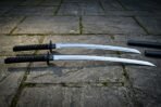 Matched katana and wakizashi daisho sword set with black cord wrapped hilts and polished steel blades resting on a flagstone courtyard beside two black lacquered scabbards