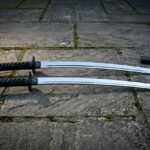 Matched katana and wakizashi daisho sword set with black cord wrapped hilts and polished steel blades resting on a flagstone courtyard beside two black lacquered scabbards