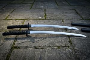 Matched katana and wakizashi daisho sword set with black cord wrapped hilts and polished steel blades resting on a flagstone courtyard beside two black lacquered scabbards
