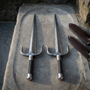 Matched pair of damascus sai daggers with upswept prongs and knurled black grips laid on a carved stone slab beside two brown leather sheaths