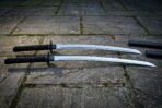 Matched pair of hand forged katana swords with Valknut engraved tsuba and black cord wrapped handles lying on grey flagstones beside two black scabbards