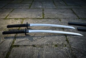 Matched pair of hand forged katana swords with Valknut engraved tsuba and black cord wrapped handles lying on grey flagstones beside two black scabbards