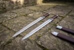 Matched Viking seax blades with brown grips angled across old paving stones, two scabbards with steel chapes beside them