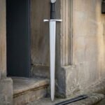 Medieval arming sword in hand forged 1095 steel with a faceted pommel and ribbed black leather grip standing in an old stone doorway