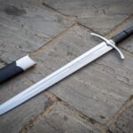 Medieval arming sword in polished high carbon steel with curved crossguard, black leather grip and wheel pommel, lying on flagstones beside a black leather scabbard
