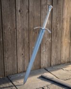 Medieval longsword in satin polished 1095 steel with an arched crossguard and knot engraved shield pommel standing upright against a weathered timber plank door