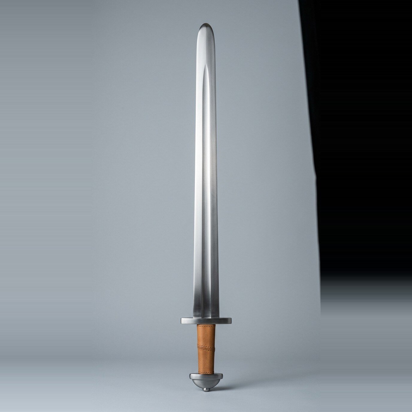 Skarnvik Migration Era Blunt Arming Sword RK-3009 Migration era arming sword with a broad shallow fuller, two part pommel and tan leather grip standing point up against a plain grey studio backdrop