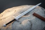 Mirror polished 1095 steel serpent etched arming sword with a knotwork engraved crossguard and lobed pommel lying across a pale sandstone boulder beside a leather scabbard