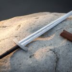 Mirror polished 1095 steel serpent etched arming sword with a knotwork engraved crossguard and lobed pommel lying across a pale sandstone boulder beside a leather scabbard