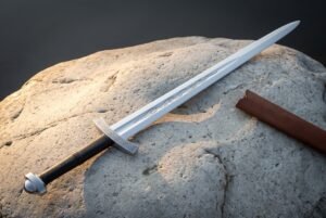 Mirror polished 1095 steel serpent etched arming sword with a knotwork engraved crossguard and lobed pommel lying across a pale sandstone boulder beside a leather scabbard