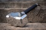 Mirror polished Mjolnir shaped steel hammer head with knotwork collar and black wrapped grip lying on a stone step