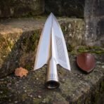 Mirror polished steel spearhead with knotwork engraved panel and runed socket standing on a mossy stone step