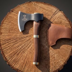 Mjolnir bearded axe with a blackened 1095 steel head and brass banded rosewood haft resting on a cut log round beside its brown leather blade cover.